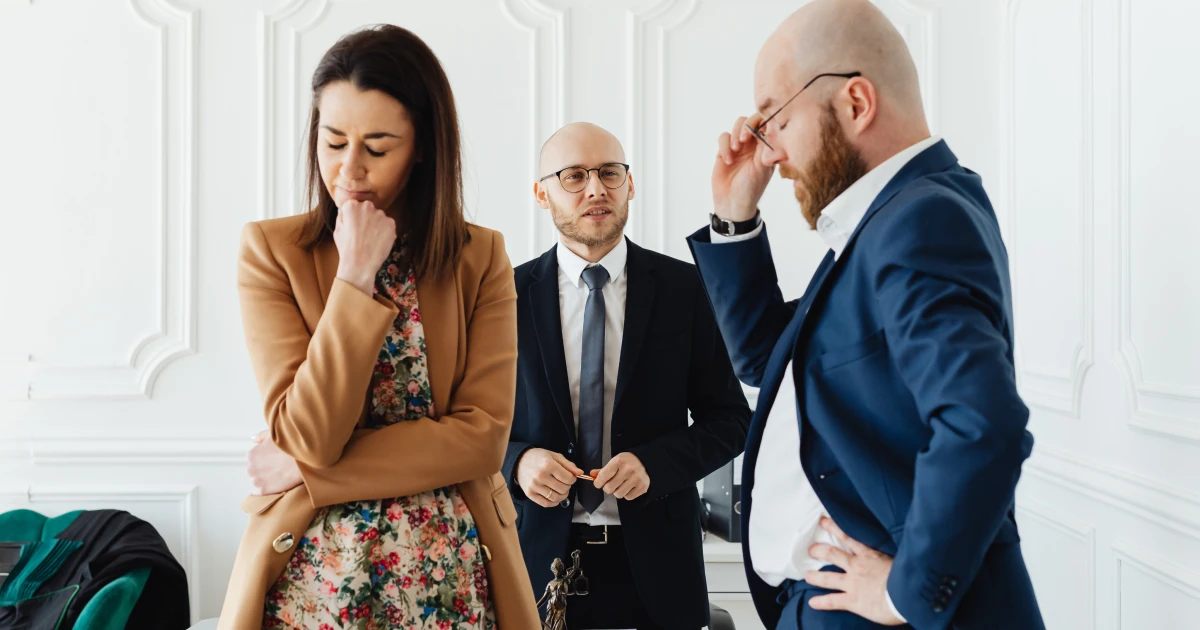 Two men in suits and a woman in a dress and coat standing in a group looking frustrated.