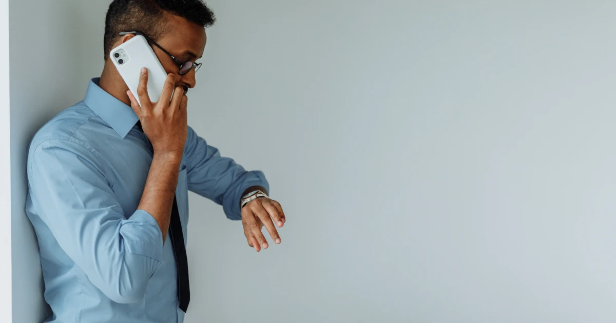 Person in button down shirt and tie standing against a wall talking on smartphone and looking at watch.