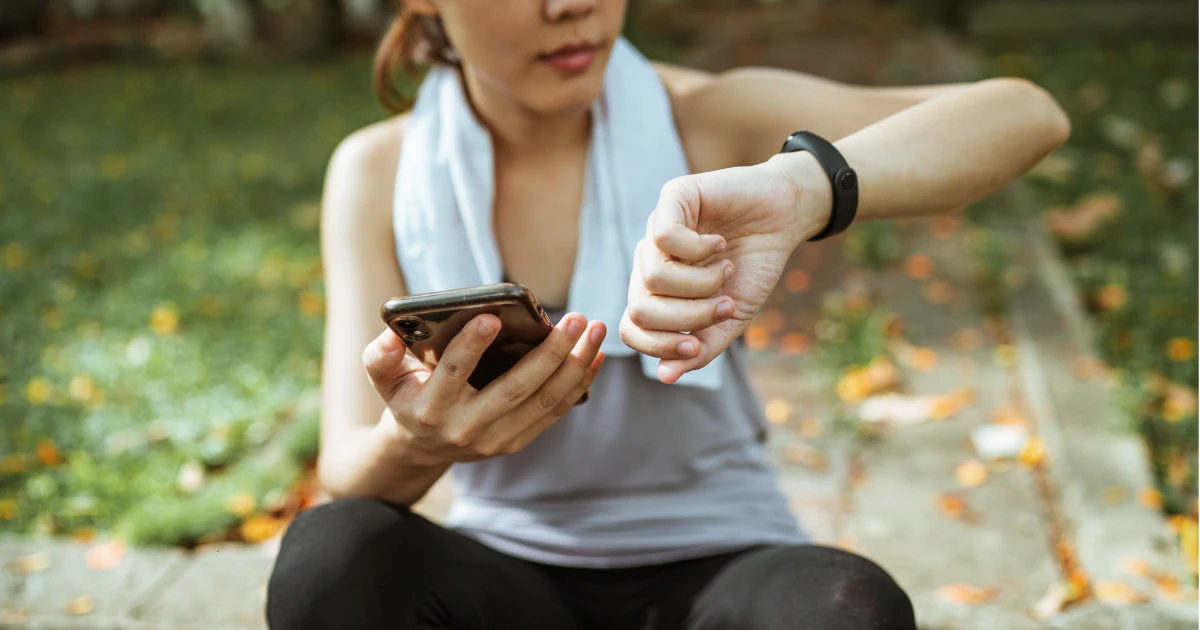 Woman sitting outside in workout clothes looking at a fitness tracker and smartphone.