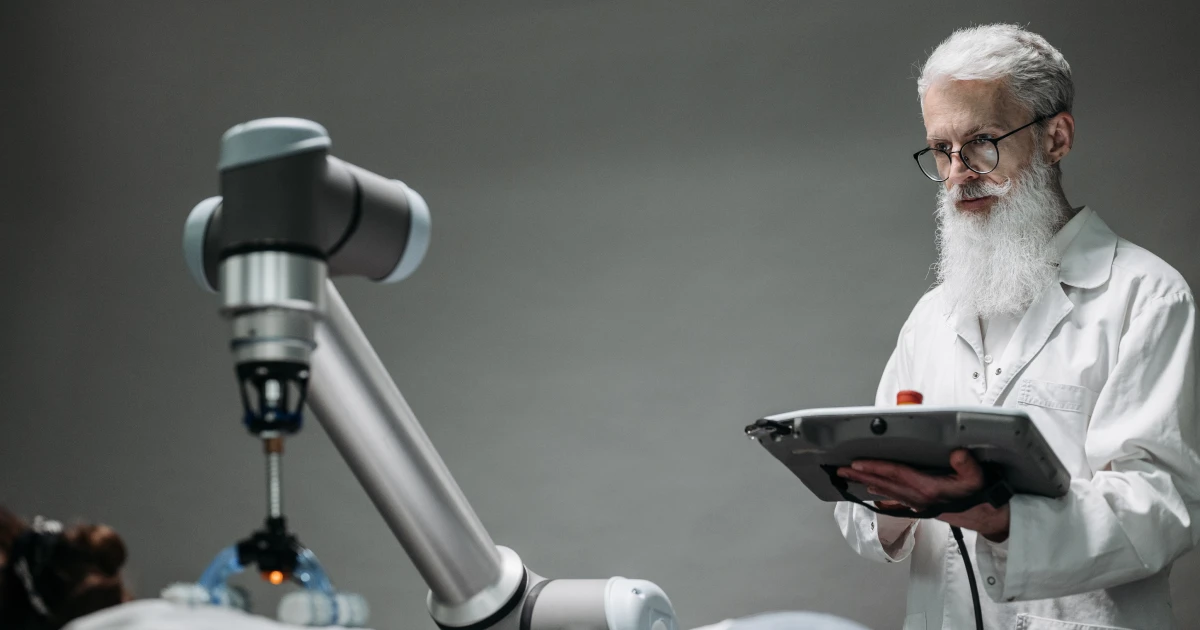 Older man in white lab coat and glasses holding control pad and watching as robotic arm performs precision surgical procedure on unseen subject.