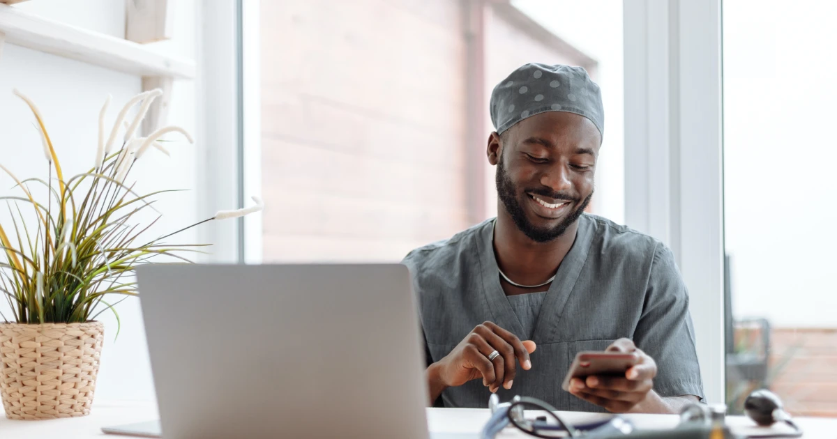 Surgeon in scrubs sitting at desk in front of open laptop looking down at smartphone and smiling.