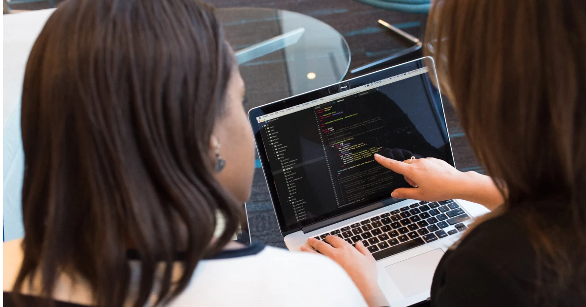 Two women looking at a computer screen displaying code, with one pointing to a line of code with her index finger.