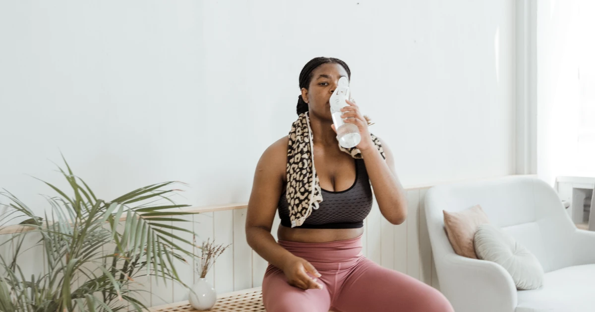 Young woman in workout gear sitting on bench in brightly lit room drinking from water bottle.