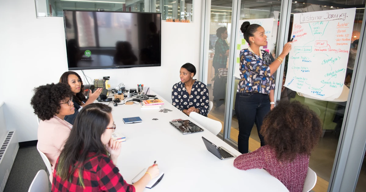Young woman leading brainstorming session in small conference room with five other team members seated around table.