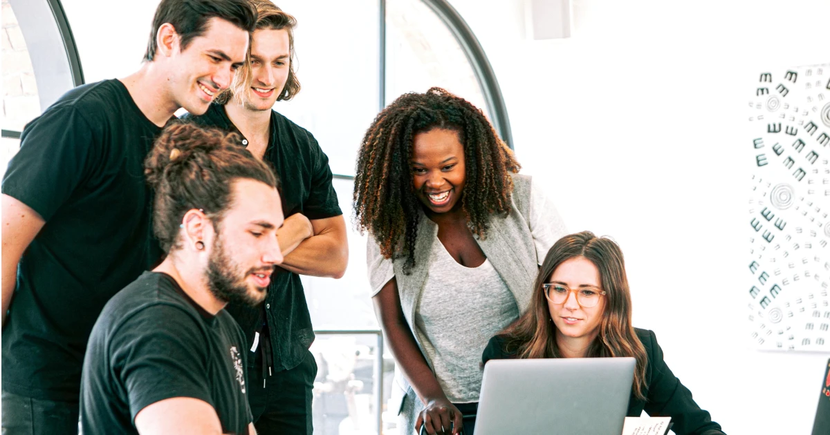 Group of people in brightly lit office standing around open laptop smiling.