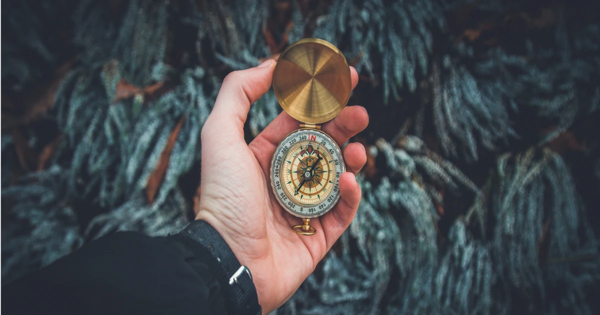 Hand holding open vintage brass compass with greenery in background.
