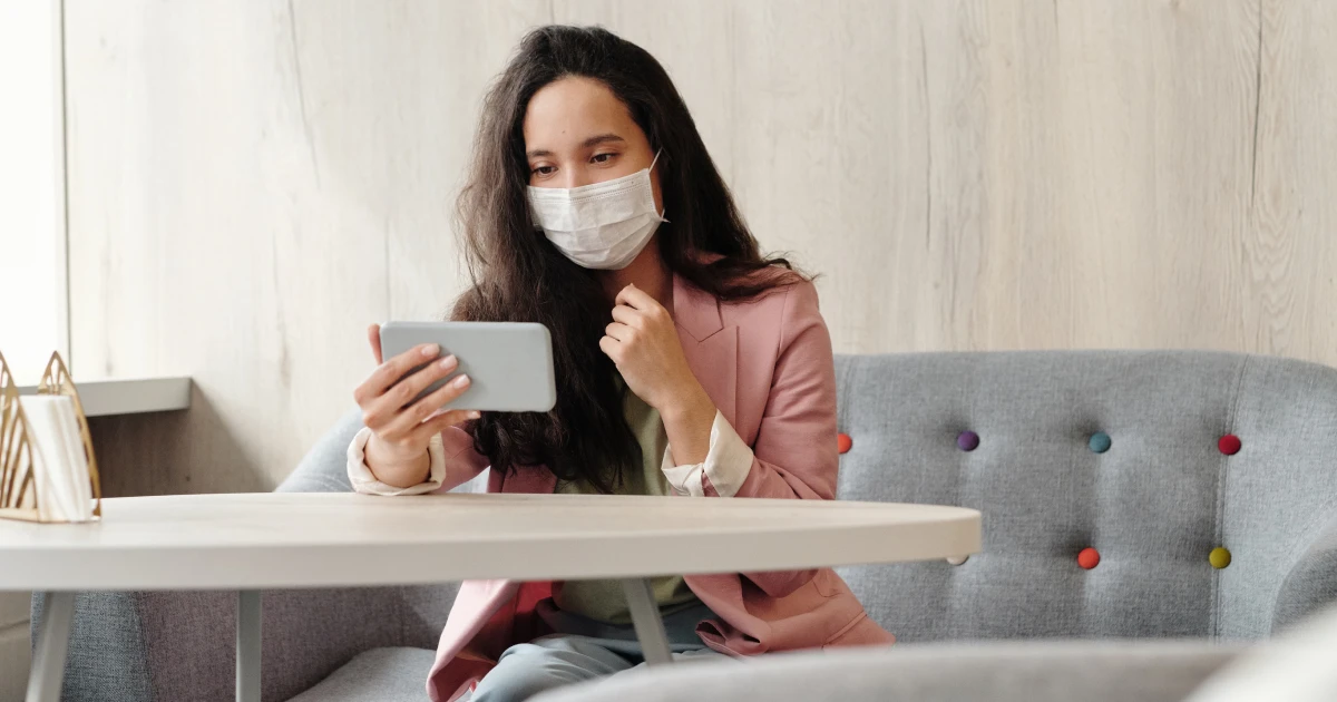 Woman sitting at booth table wearing surgical mask having video chat on smart phone.