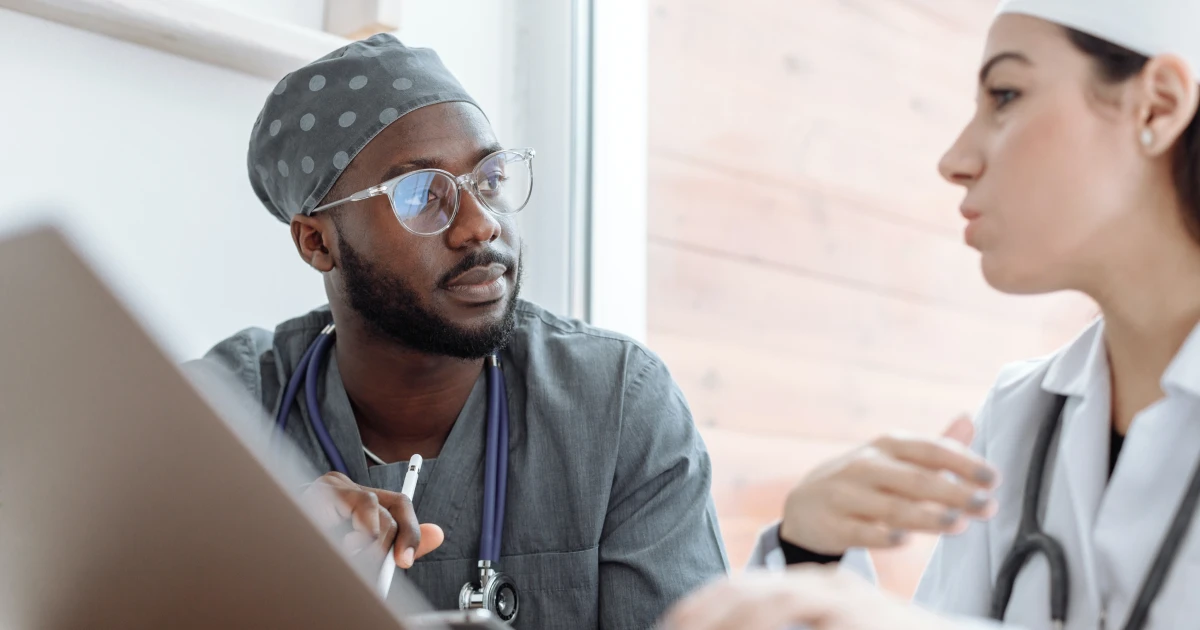 Two surgeons in scrubs talking while referring to info on open laptop.