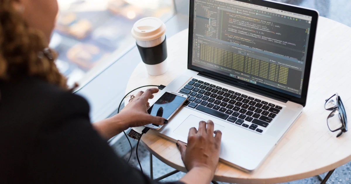 Woman using open laptop displaying code with smartphone sitting on top, and coffee and glasses on table beside it.